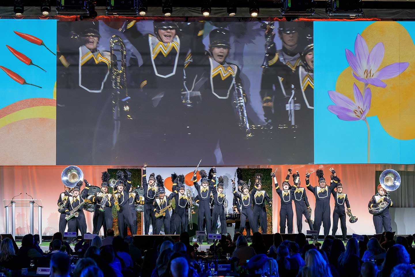 A marching band performing on stage at the Global Produce and Floral show with large screen and colorful graphics in the background.