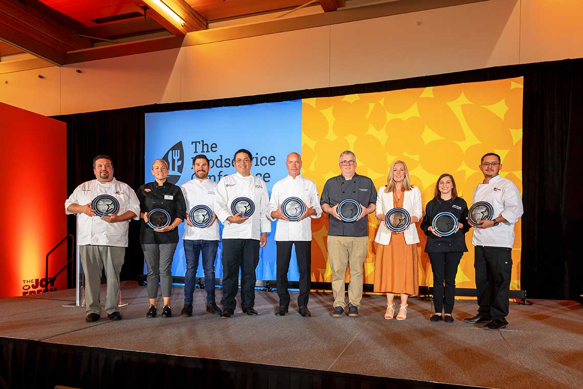 Nine people standing on a stage holding plates, smiling. The backdrop features a blue and orange display with the text "The Foodservice Conference." The group appears to be diverse, including men and women, dressed in business casual and chef attire. Nine people standing on a stage holding plates, smiling. The backdrop features a blue and orange display with the text "The Foodservice Conference." The group appears to be diverse, including men and women, dressed in business casual and chef attire.