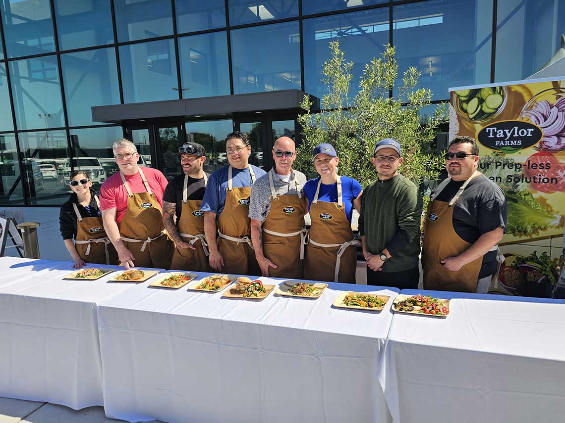 A group of eight people dressed in aprons and casual clothing stand behind a table displaying various plated dishes. They are posed outdoors in front of a modern glass building. A banner for Taylor Farms is visible in the background. A group of eight people dressed in aprons and casual clothing stand behind a table displaying various plated dishes. They are posed outdoors in front of a modern glass building. A banner for Taylor Farms is visible in the background.