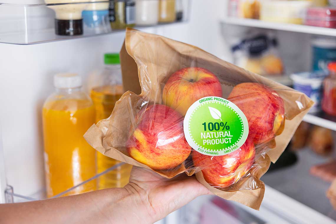 A hand holding a packaged set of apples in front of an open refrigerator.