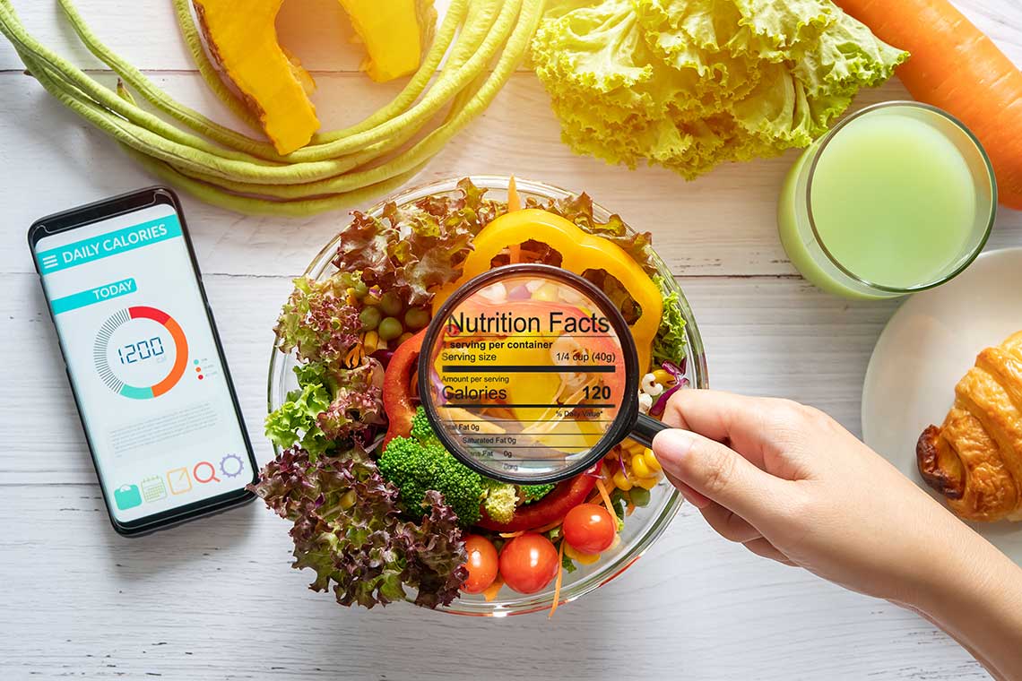 A bowl of salad with a magnifying glass showing nutrition facts, surrounded by a smartphone, croissant, glass of juice, and fresh vegetables.