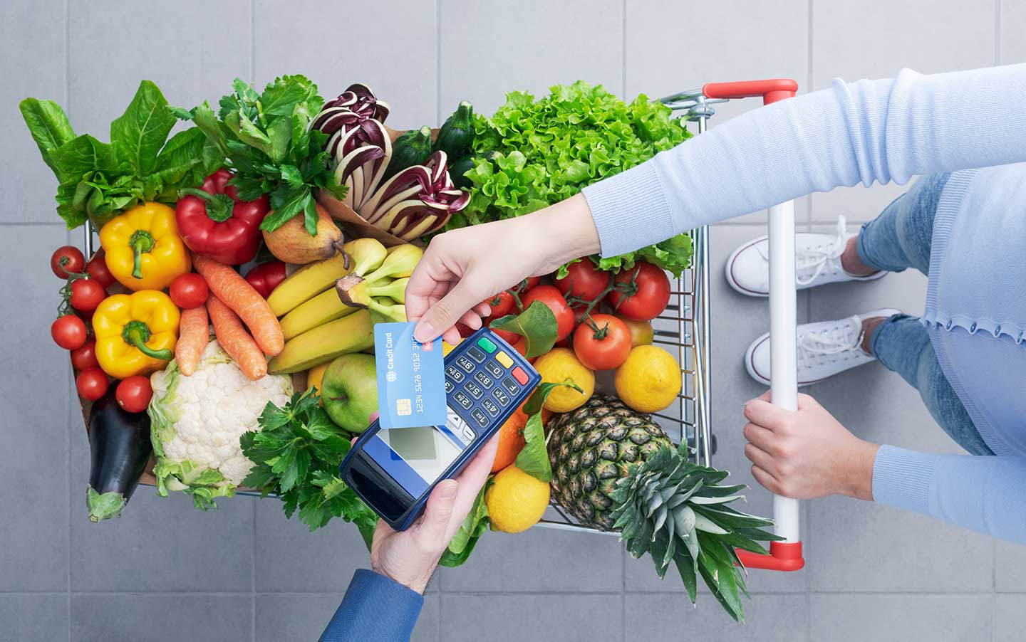 Woman pushing a shopping cart full of fresh vegetables and fruits, she is holding a delicious apple, top view