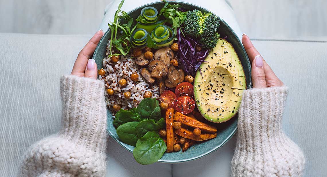 A colorful bowl of vegetables and quinoa held by hands with cream knit sleeves.