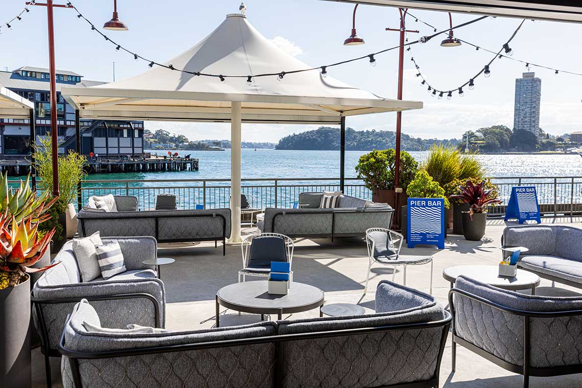 Outdoor seating area with sofas and tables under a white canopy by the water.