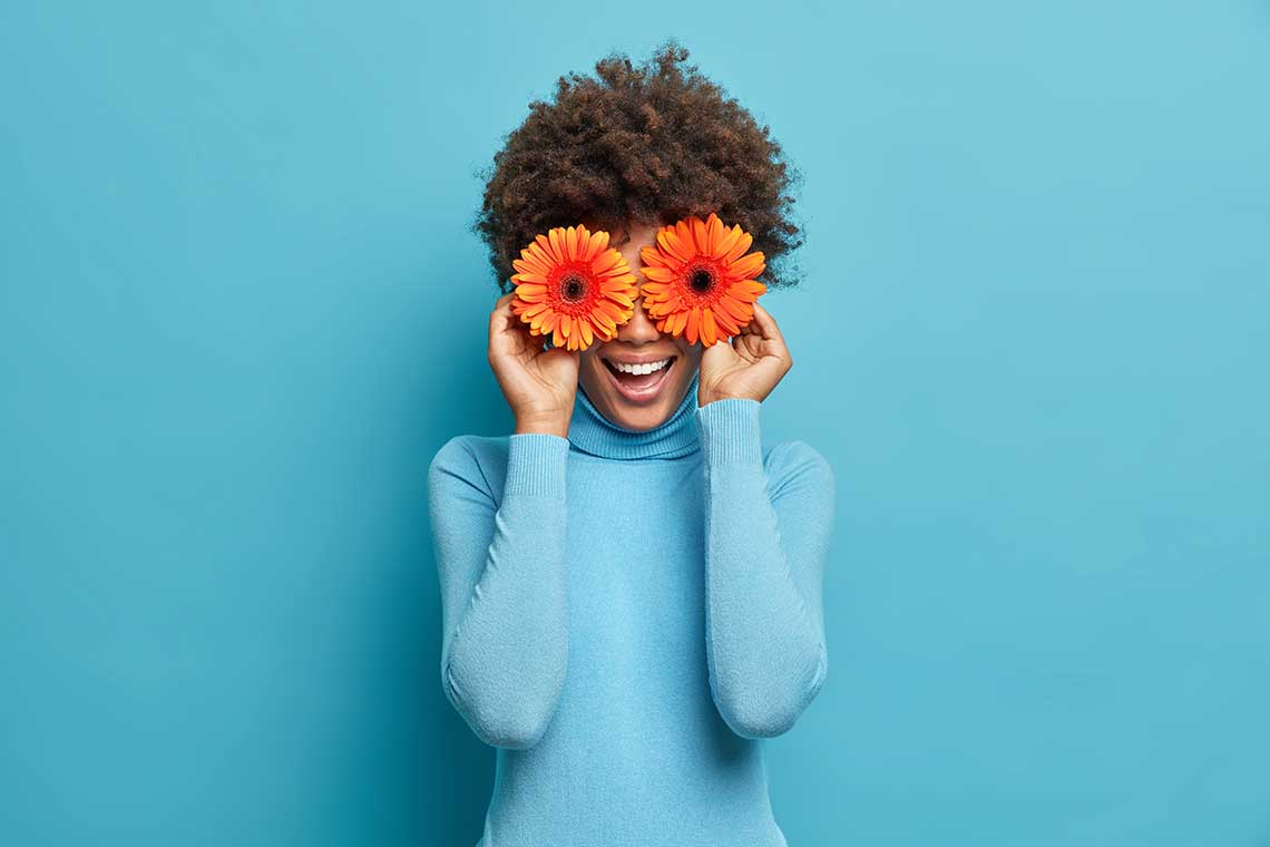 Person holding orange flowers over eyes, smiling against a blue background.