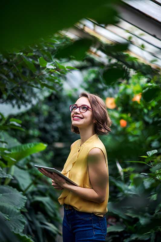 A woman in a yellow blouse and glasses, holding a tablet, smiles while standing in a lush greenhouse.