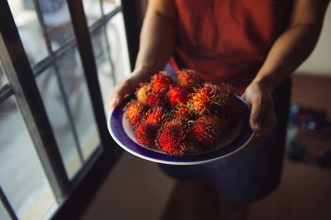 Hands holding a plate of red rambutan fruits by a window.
